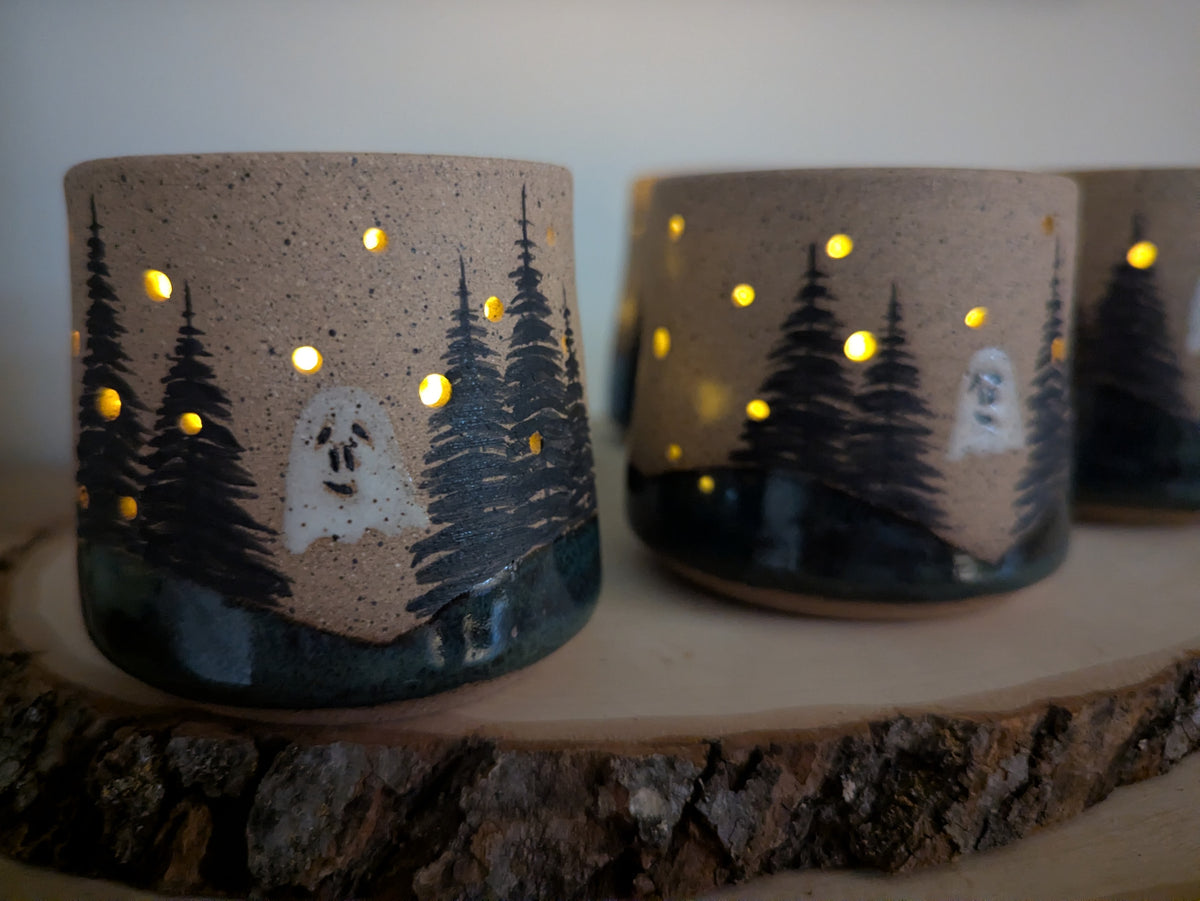 Two ceramic candle holders with a forest and ghost design, lit from within to cast warm yellow light through the cutouts, displayed on a rustic wood slice.