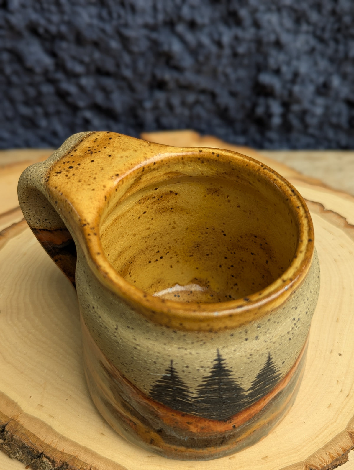 A handmade ceramic mug with a handle, featuring a rustic glaze in earthy brown, beige, and orange tones, with a black pine tree design near the base. The mug is displayed on a natural wood slice against a dark textured background.
