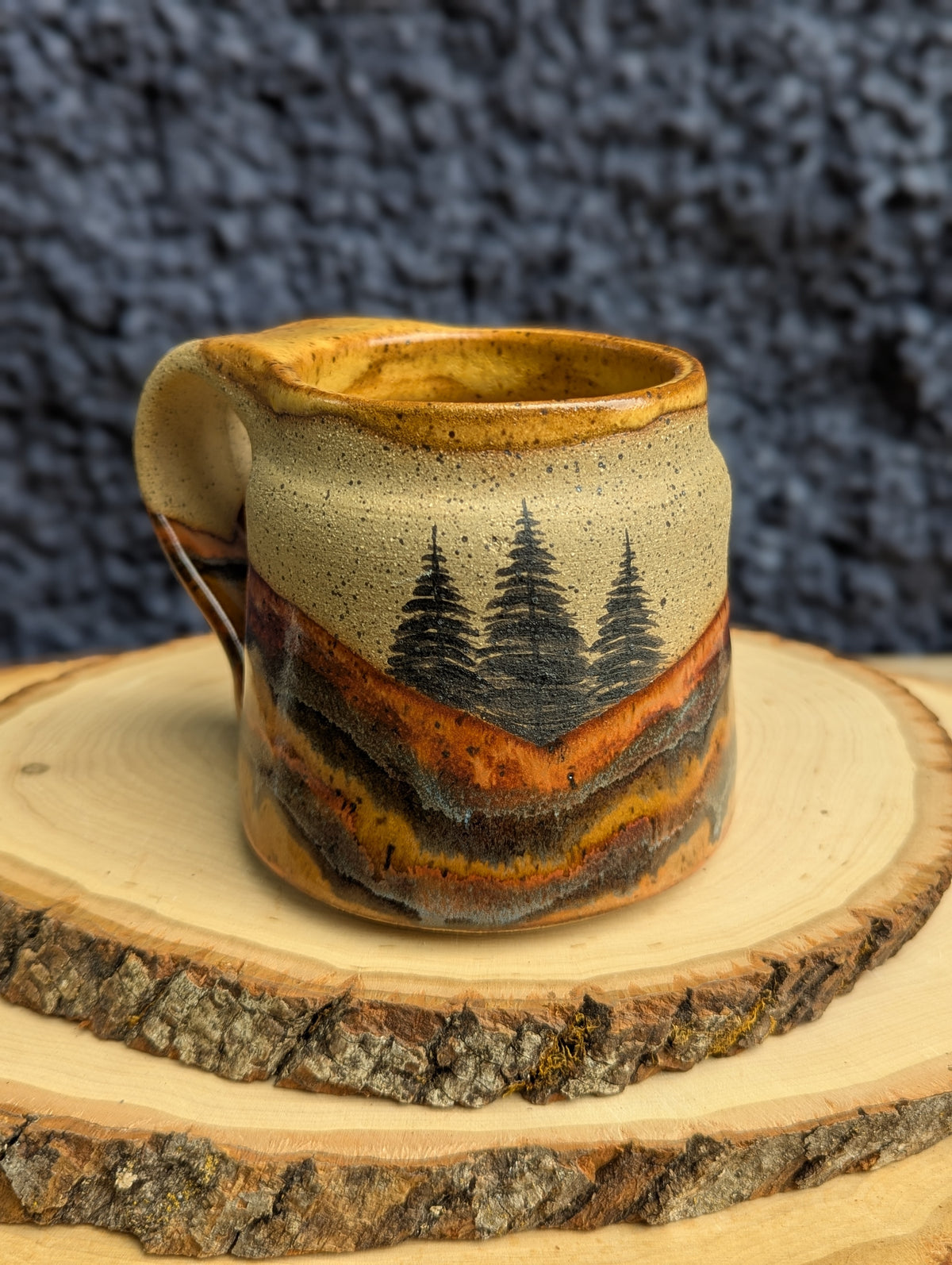 A handmade ceramic milk pitcher with a handle, featuring a rustic landscape design of pine trees and rolling hills in earthy brown, orange, and beige glazes. The pitcher is displayed on a rustic wood slice against a dark textured background.
