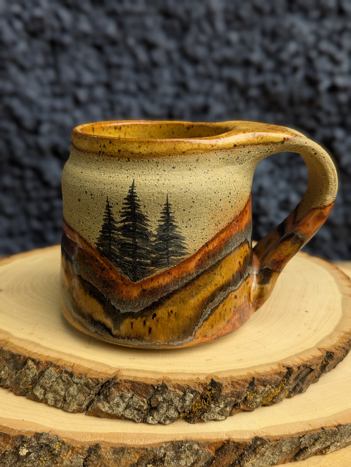 A handmade ceramic mug with a rustic hand-painted landscape design of pine trees and rolling hills in earthy brown, orange, and beige tones, displayed on a wood slice against a dark textured background.