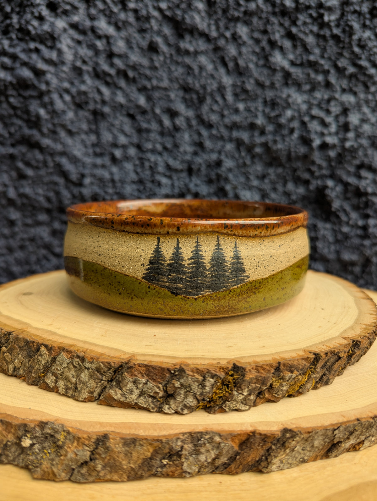 A handmade ceramic bowl with a natural wood-grain rim and a landscape design of pine trees on a hillside, displayed on a rustic wood slice against a dark textured background.