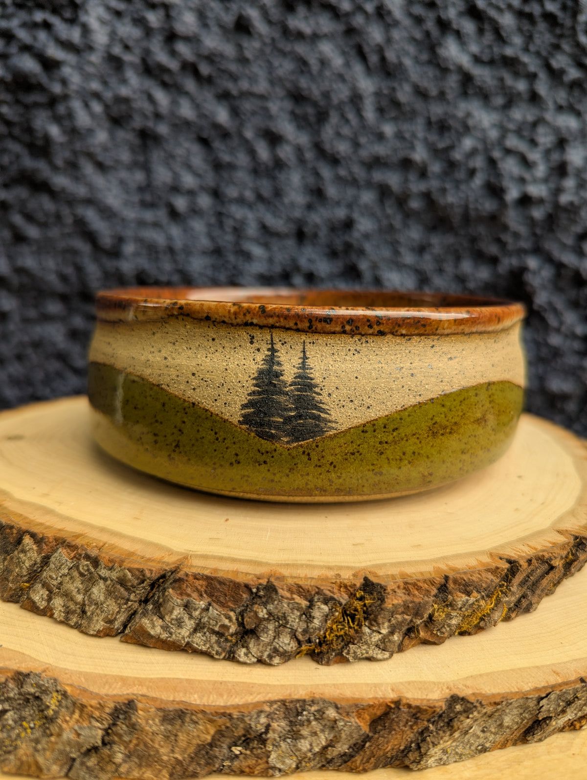 A handmade ceramic bowl with a rustic landscape design, featuring a brown rim, a beige upper section, and a green lower section with two black pine tree silhouettes. The bowl is displayed on a rustic wood slice against a dark textured background.