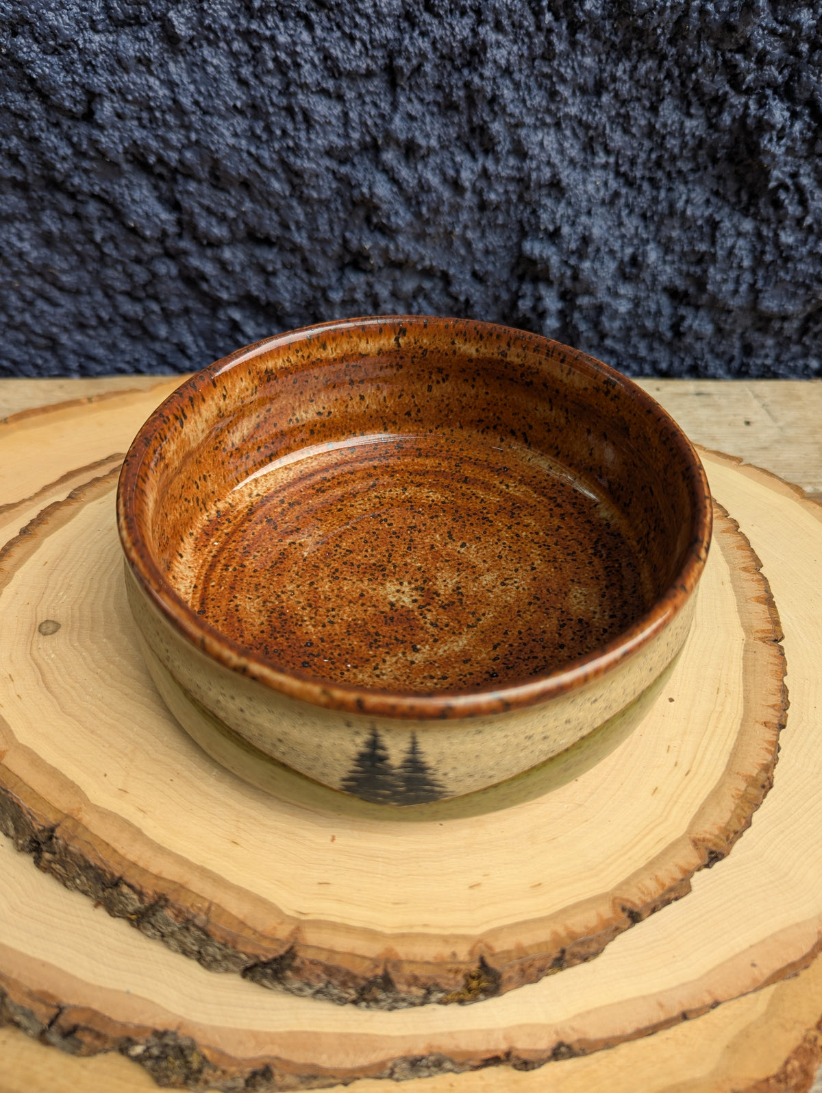 A handmade ceramic bowl with a rustic brown and amber glaze, displayed on a stacked wood slice coaster against a dark textured background.