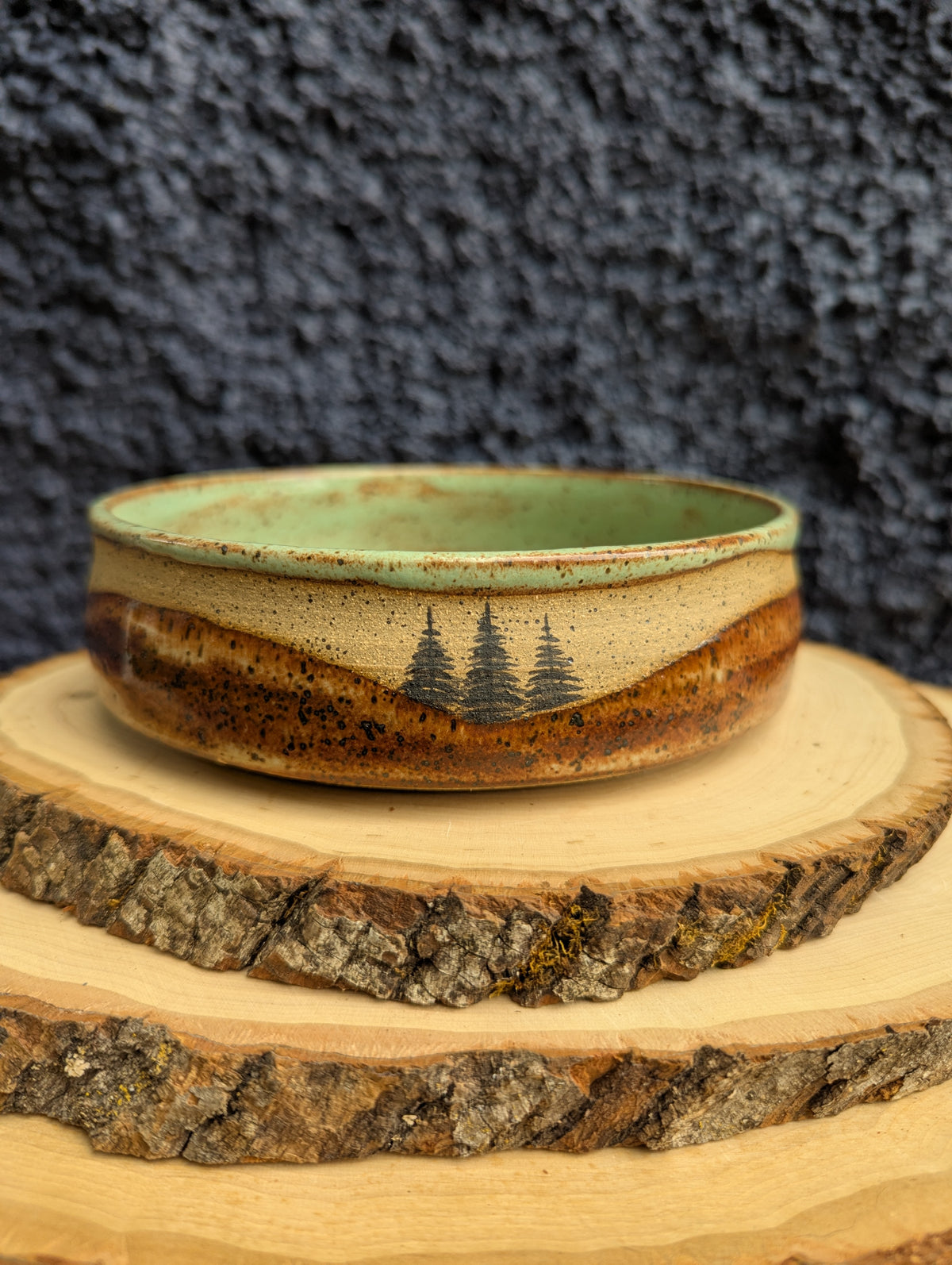 A handmade ceramic bowl with a natural wood-grain pattern and three pine tree illustrations, displayed on rustic wood slices against a dark textured background.