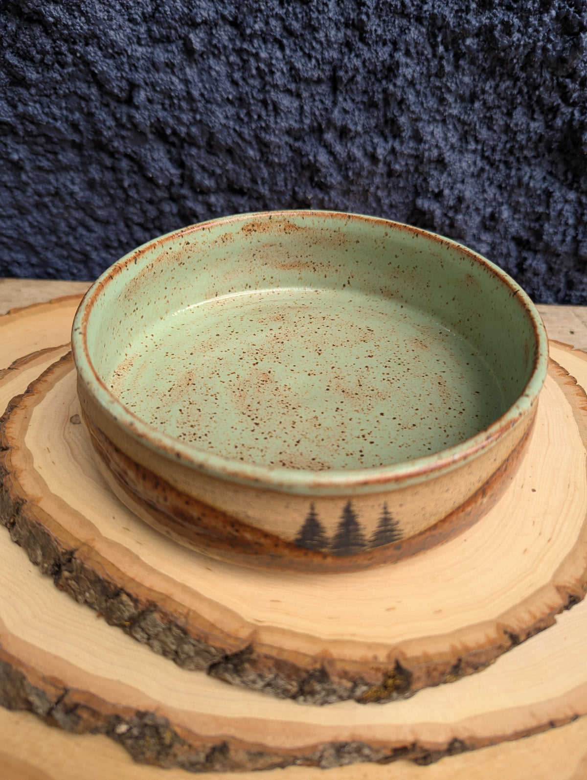 A handmade ceramic bowl with a speckled sage green interior and a rustic brown exterior featuring a small black tree design, displayed on a wood slice coaster against a dark textured background.