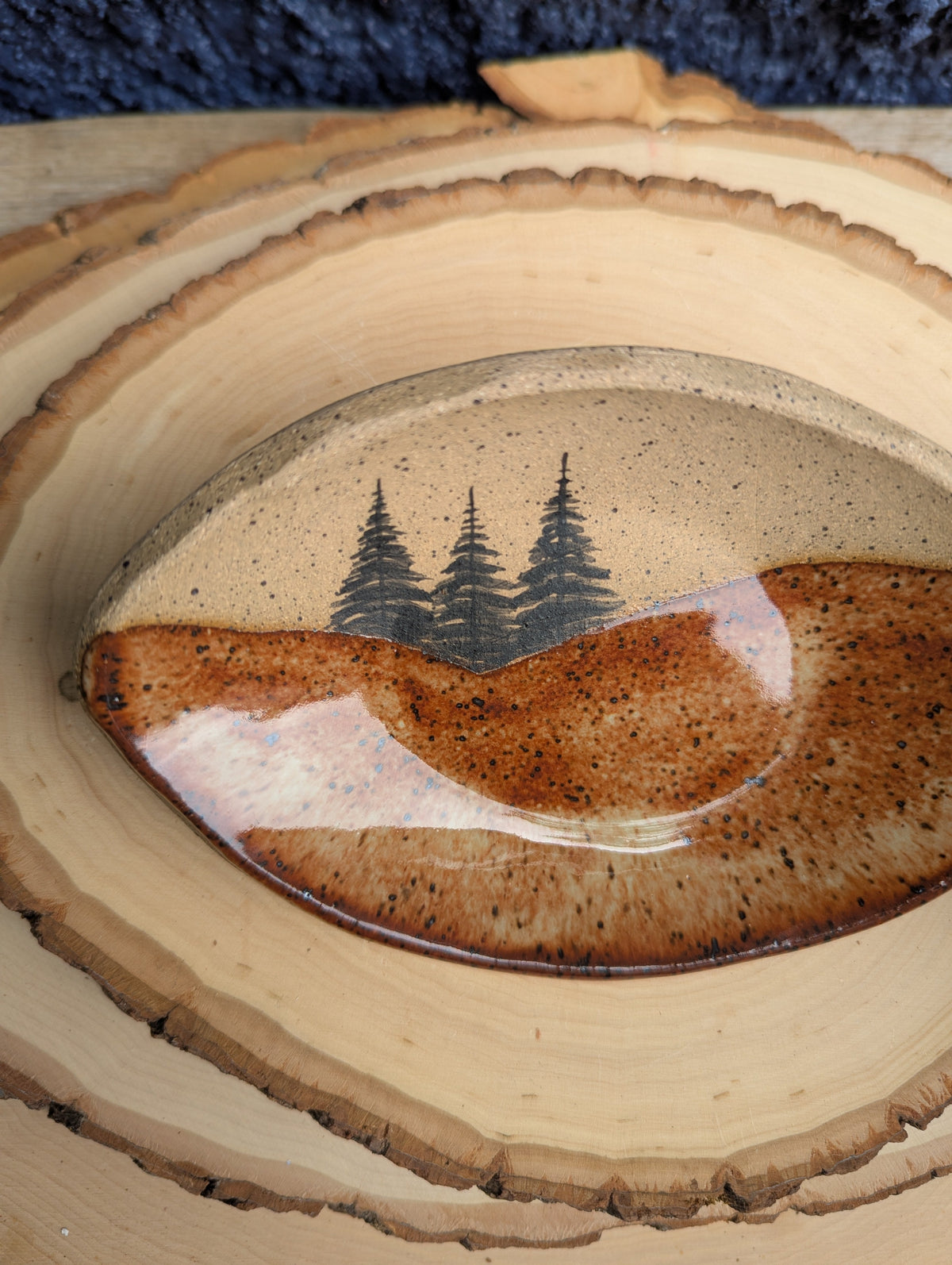 A close-up of a handcrafted wooden bowl or tray with a natural wood slice edge, featuring a glossy brown resin inlay with a black pine tree design and a small pink accent.