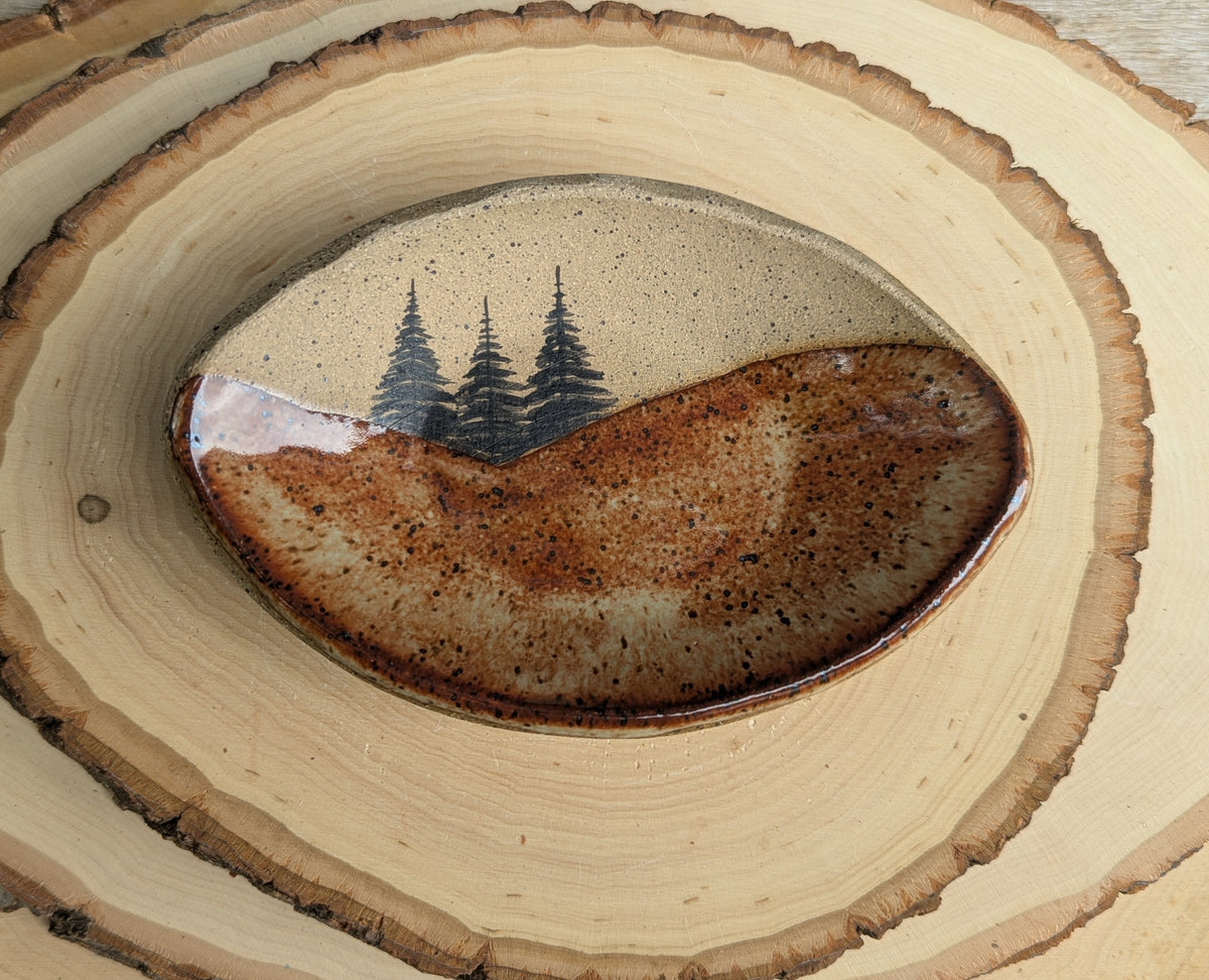 A rustic ceramic bowl with a glossy brown and beige landscape design featuring three pine trees, set on a stack of natural wood slices.