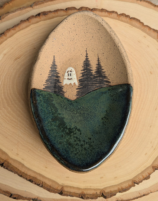A handmade ceramic dish with a rustic design. The top half is a speckled tan glaze featuring a hand-painted scene of three dark pine trees and a small white ghost.