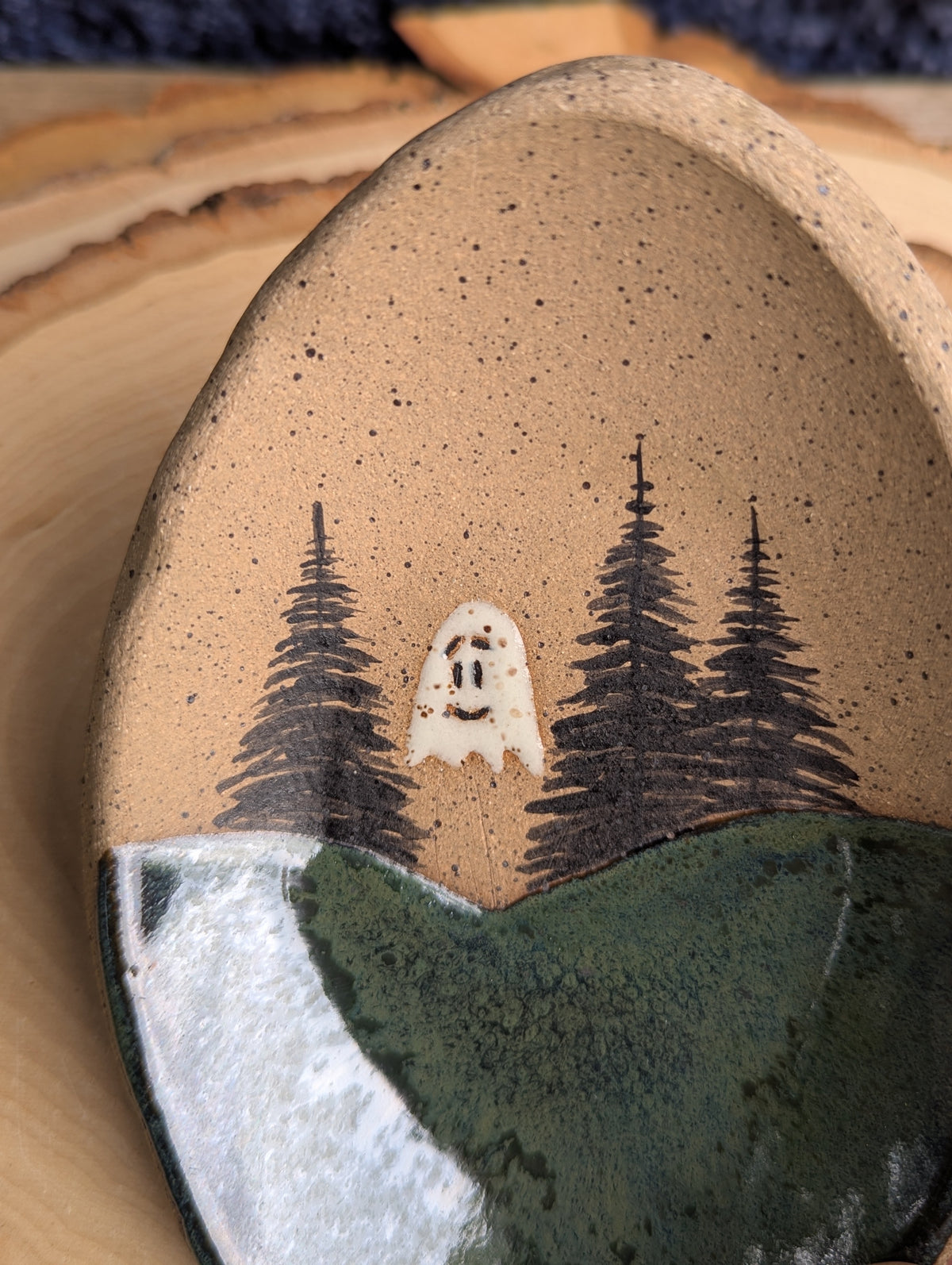 A close-up of a handmade ceramic dish with a rustic design. The top half is a speckled tan glaze featuring a hand-painted scene of three dark pine trees and a small white ghost.