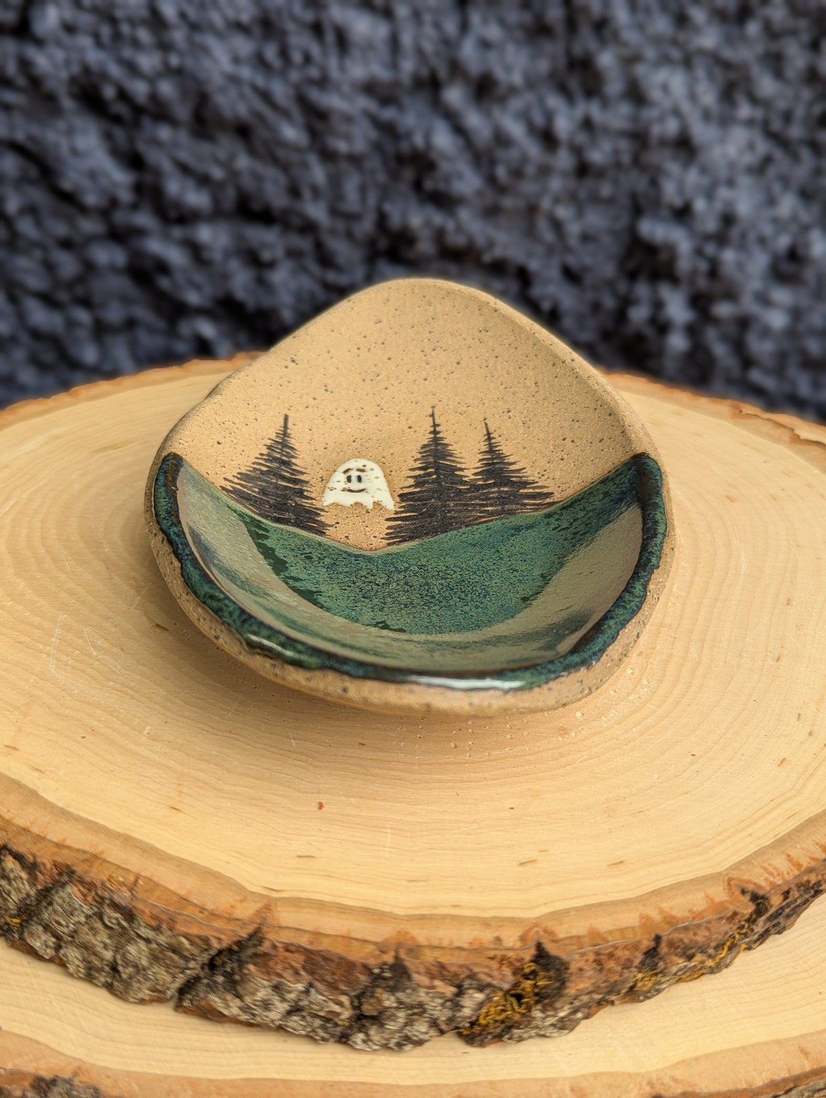A small handmade ceramic dish with a hand-painted forest landscape design, displayed on a rustic wood slice against a dark textured background.
