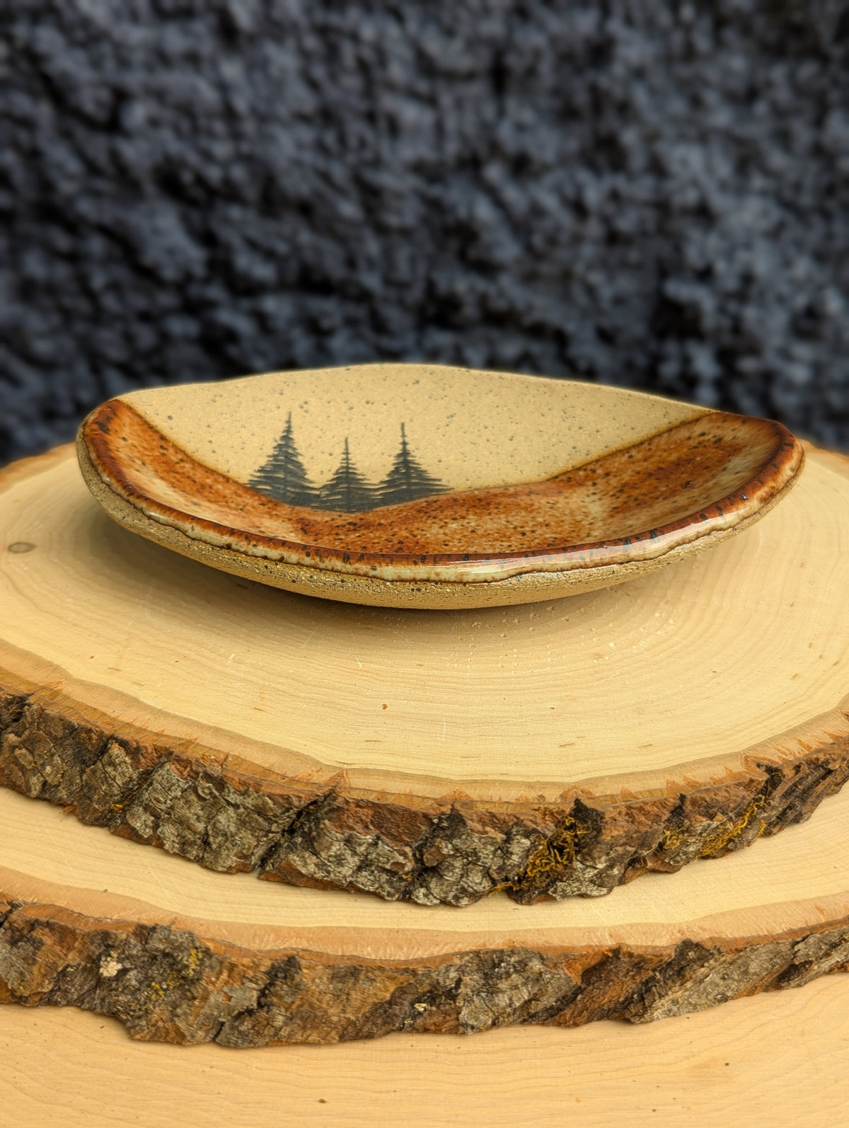 A handmade ceramic dish with a natural wood-grain rim and a painted pine tree design, displayed on stacked wood slices against a dark textured background.