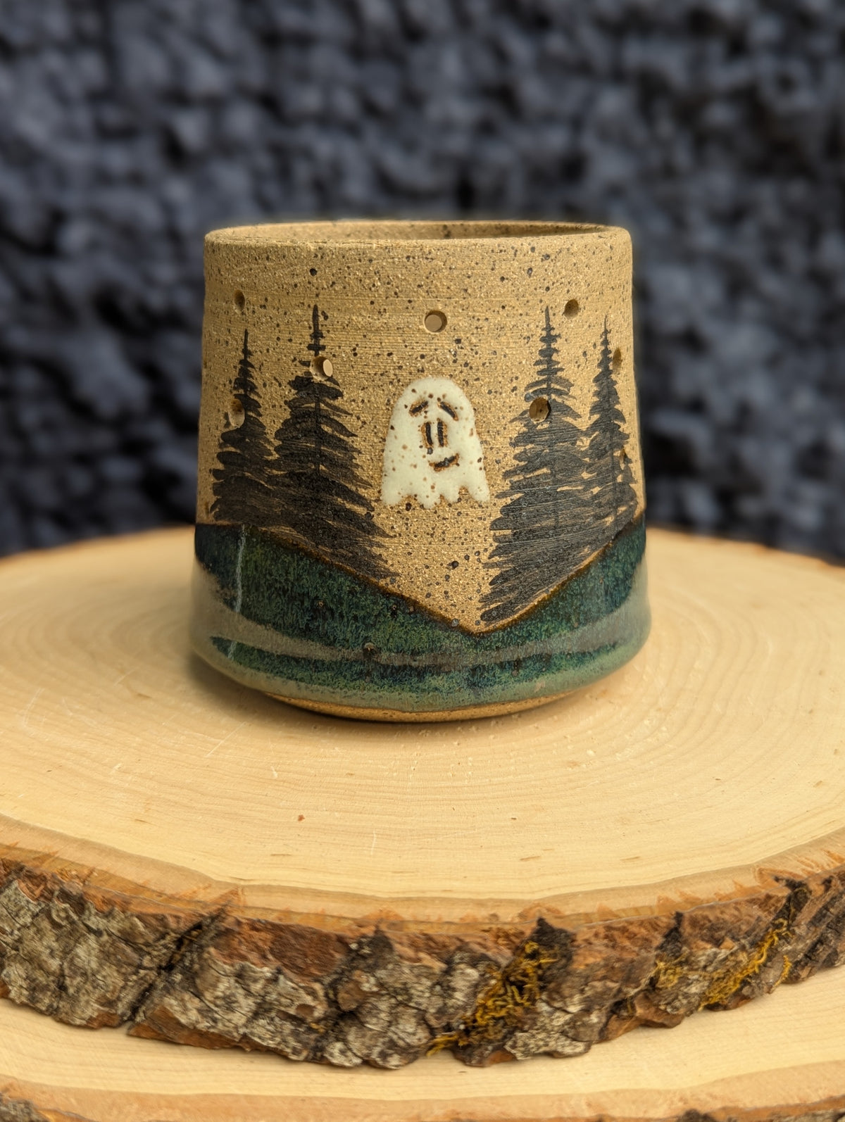 A handmade ceramic cup featuring a forest landscape design with pine trees and a white ghost, displayed on a rustic wood slice against a dark textured background.