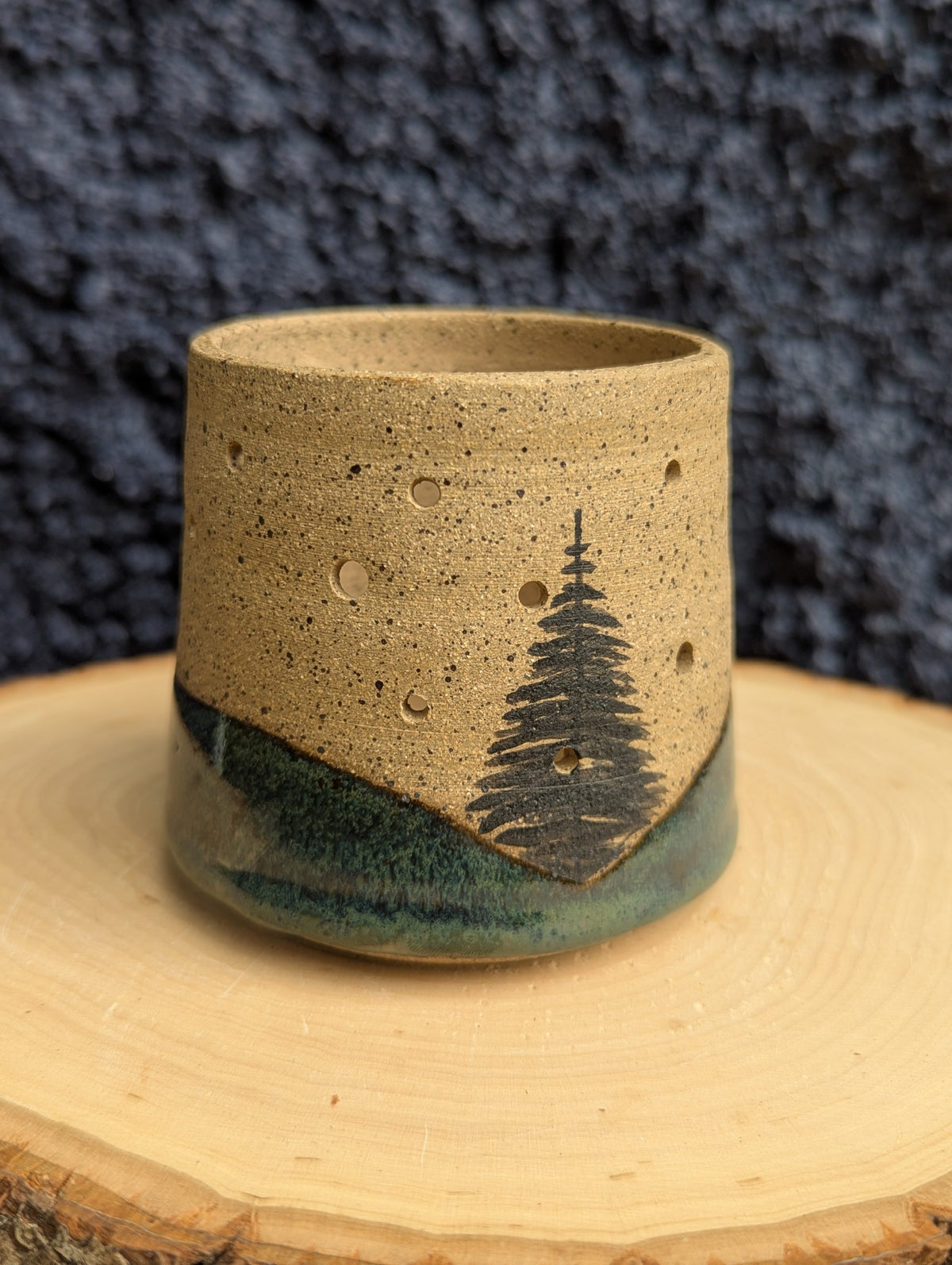 A handmade ceramic cup with a speckled beige upper body, a dark green glazed lower section, and a black painted pine tree design, displayed on a wooden slice against a dark textured background.