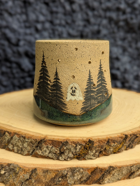 A handmade ceramic cup with a natural sandy beige glaze, decorated with a hand-painted forest scene featuring pine trees, a green hill, and a white moon. The cup is displayed on a rustic wood slice against a dark textured background.
