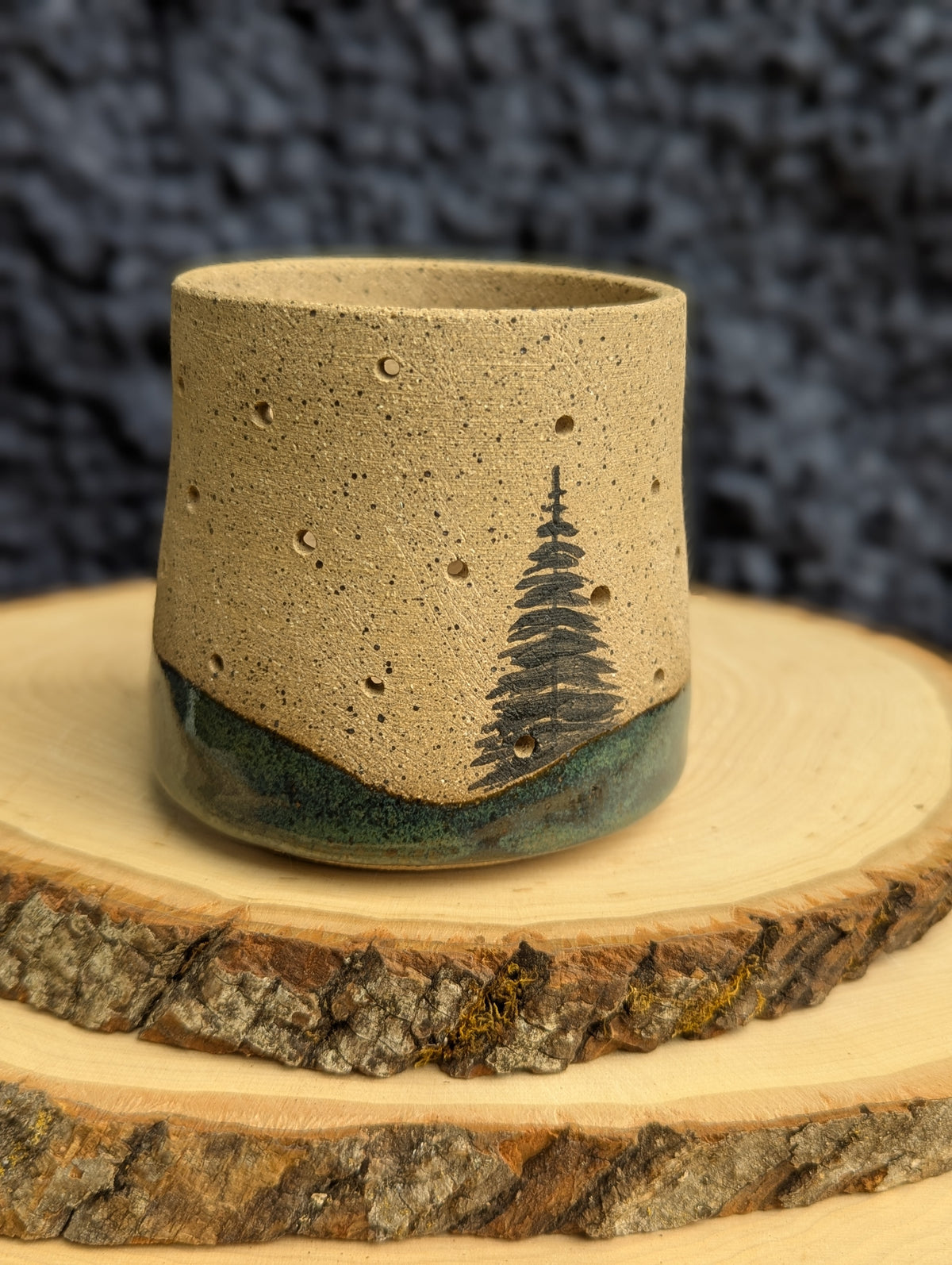 A handmade ceramic cup with a speckled beige top half and a glossy dark green base, decorated with a black pine tree design. The cup is displayed on a rustic wood slice against a dark textured background.