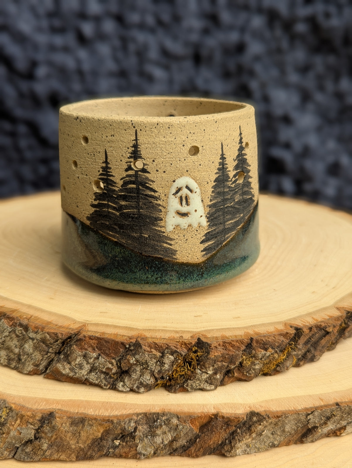 A handmade ceramic cup with a forest and ghost design, displayed on a rustic wood slice against a dark textured background.