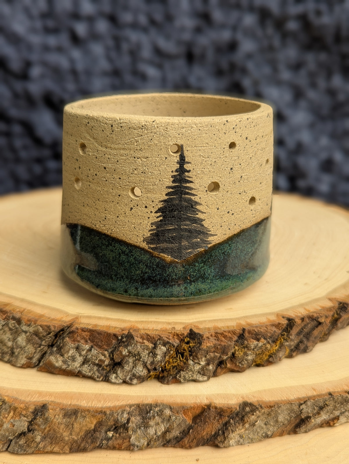 A handmade ceramic ring with a textured beige top half featuring a black pine tree design and small perforations, and a dark green glazed bottom half, displayed on a rustic wood slice against a dark textured background.