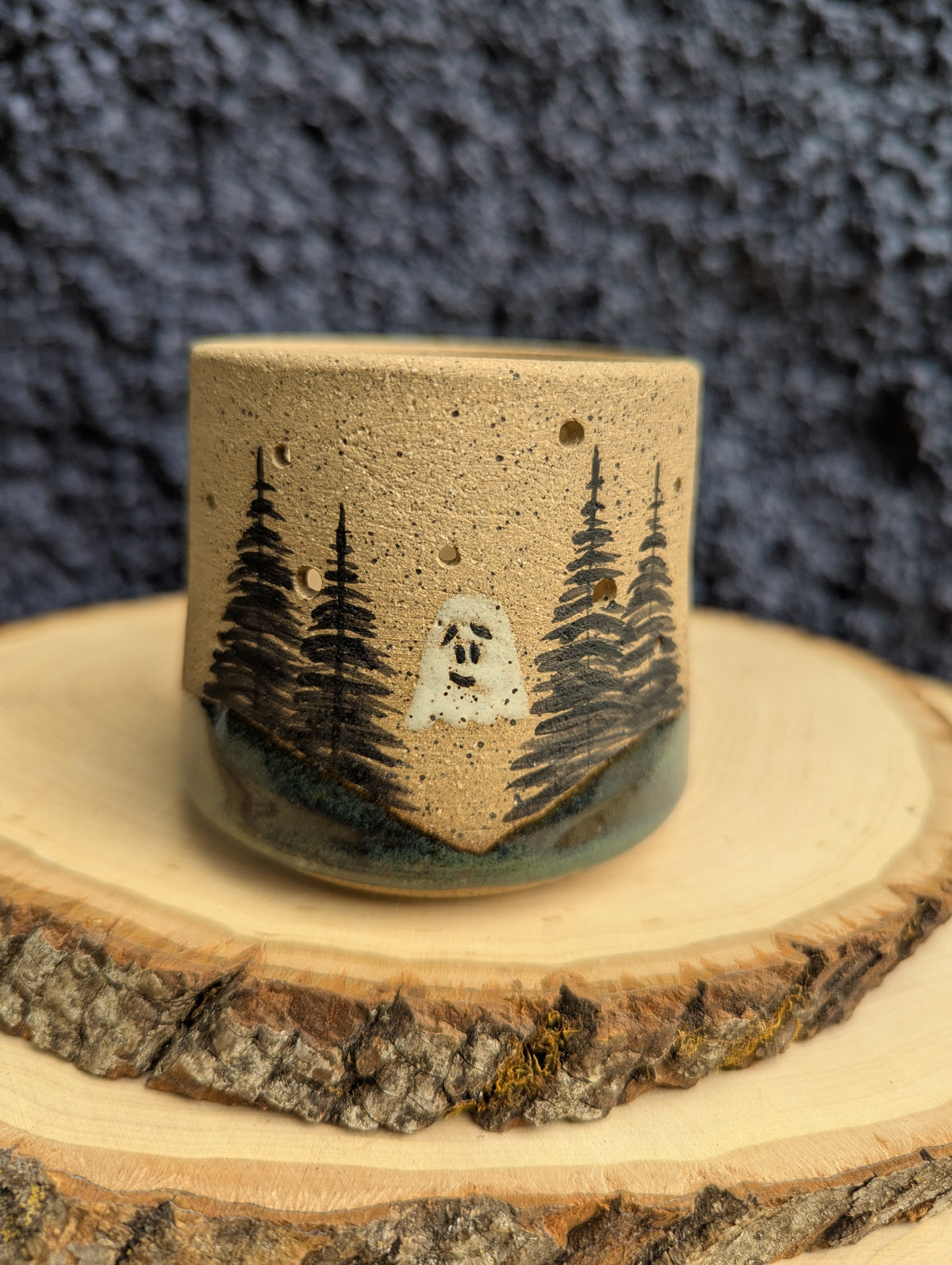 A handmade ceramic cup with a natural sandy beige body, decorated with black painted pine trees and a small white ghost, resting on a rustic wood slice against a dark textured background.
