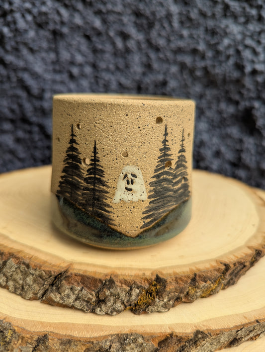 A handmade ceramic cup with a natural sandy beige body, decorated with black painted pine trees and a small white ghost, resting on a rustic wood slice against a dark textured background.