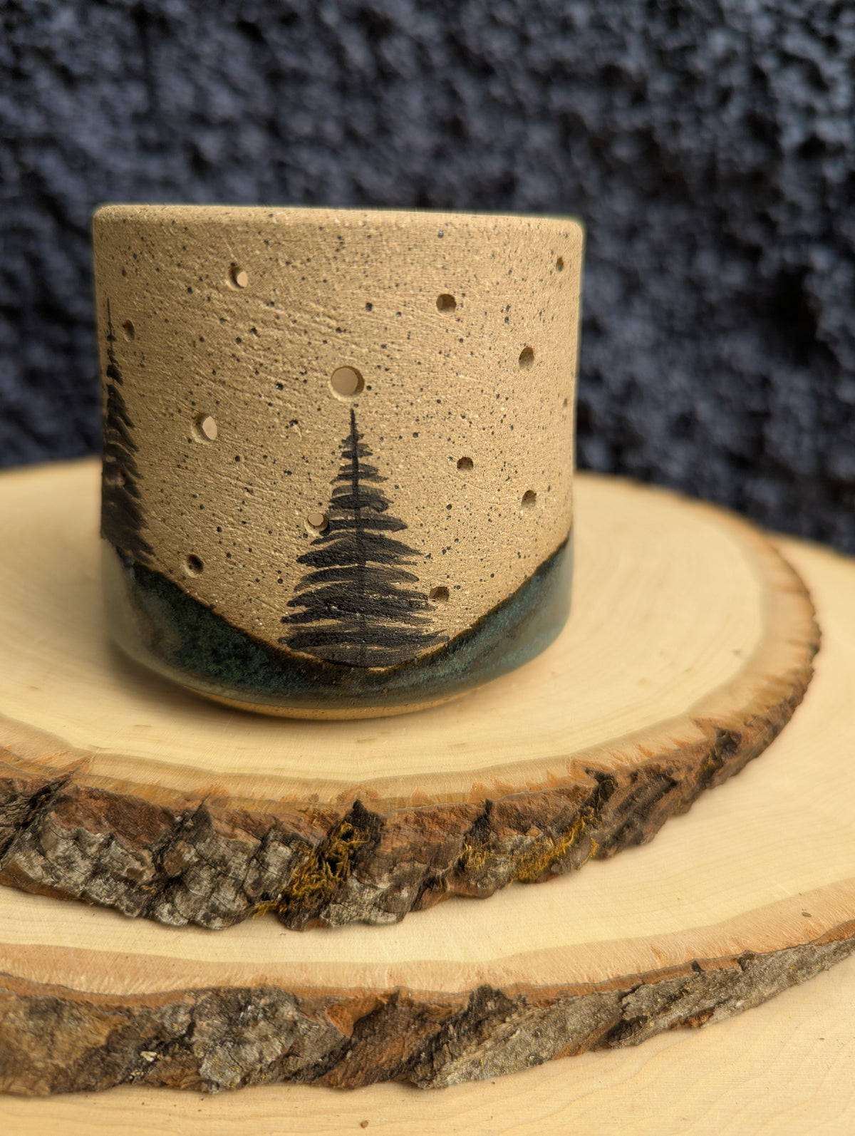 A handmade ceramic cup with a natural sandy beige body, featuring a hand-painted black pine tree design and a dark green glazed base, displayed on rustic wood slices against a dark textured background.
