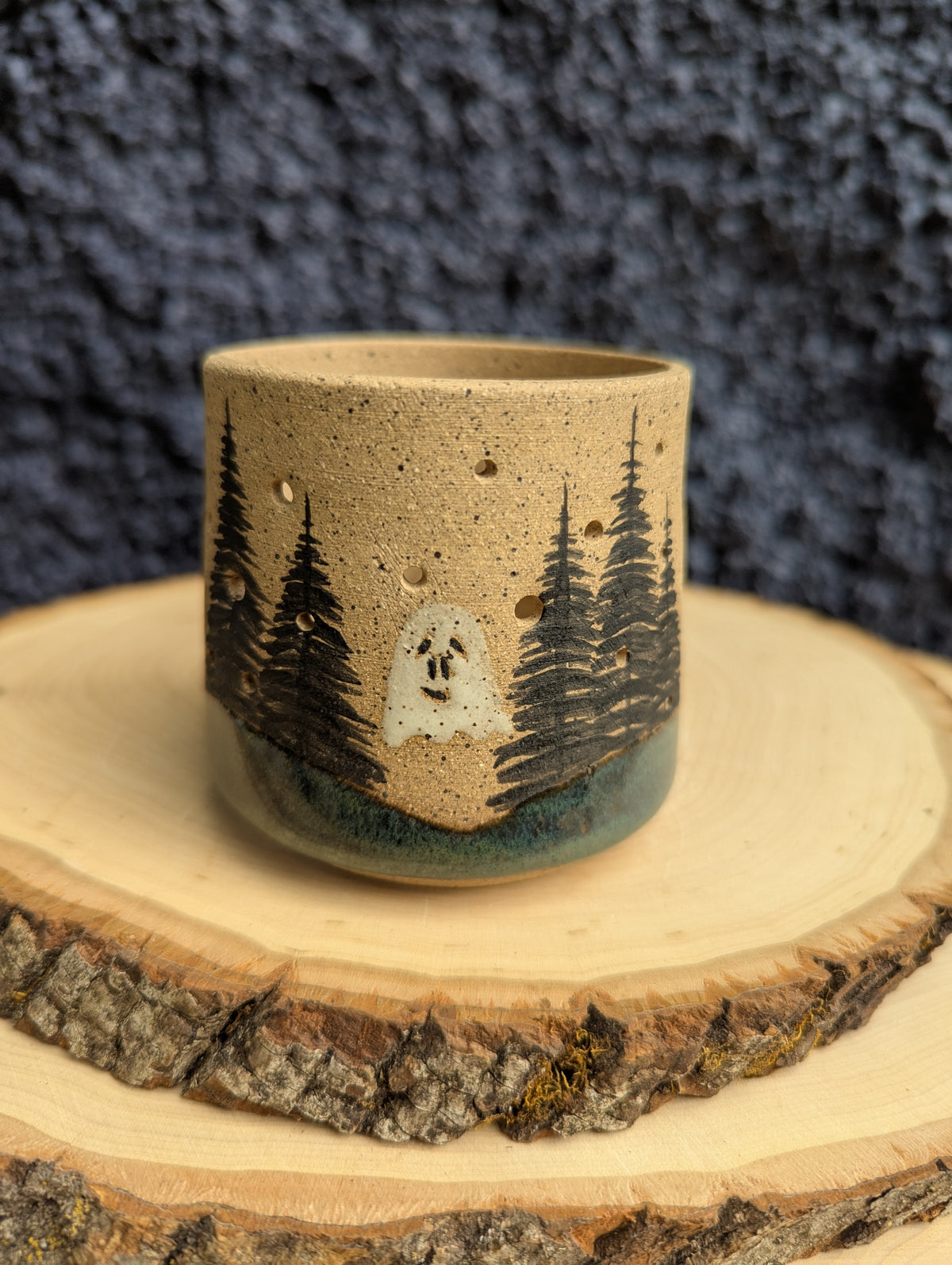 A handmade ceramic cup with a rustic design, featuring a hand-painted ghost and pine trees on a speckled beige clay body, resting on a wood slice against a dark textured background.
