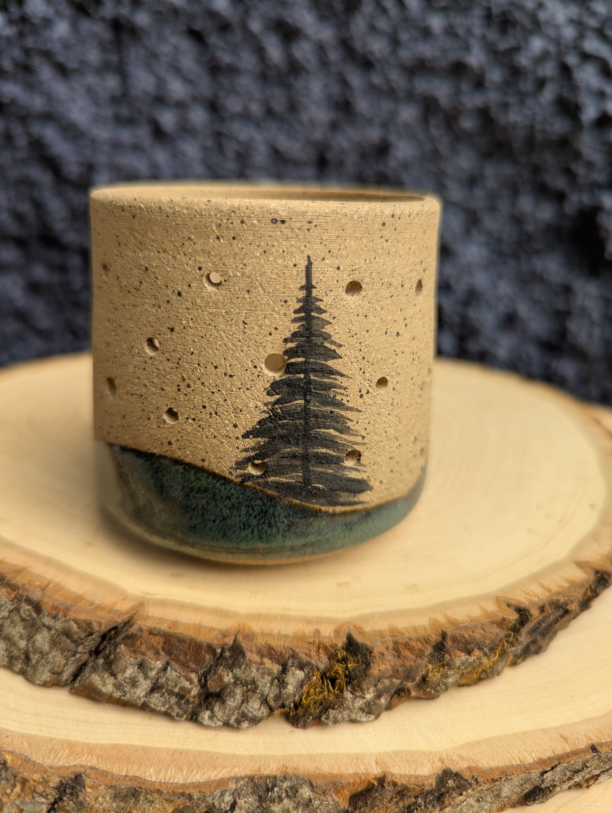 A handmade ceramic cup with a speckled beige top and a dark green glazed base, featuring a black tree design on the front, displayed on a rustic wood slice against a dark textured background.