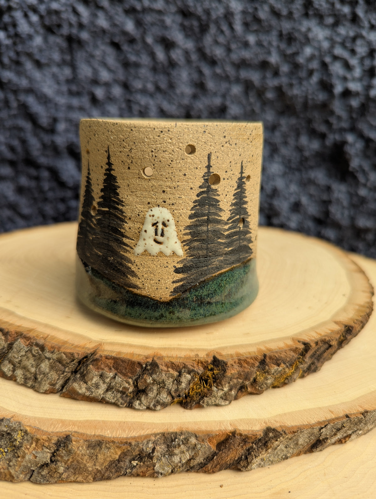 A handmade speckled ceramic cup with a forest and ghost design, displayed on a rustic wood slice against a dark textured background.
