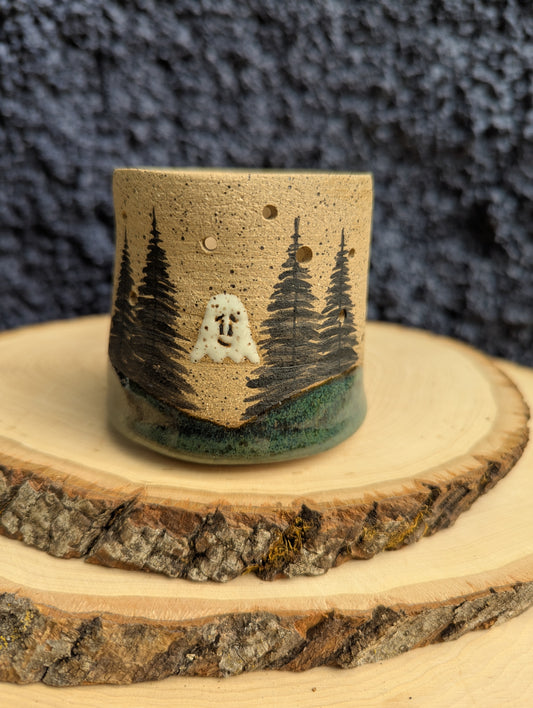 A handmade speckled ceramic cup with a forest and ghost design, displayed on a rustic wood slice against a dark textured background.
