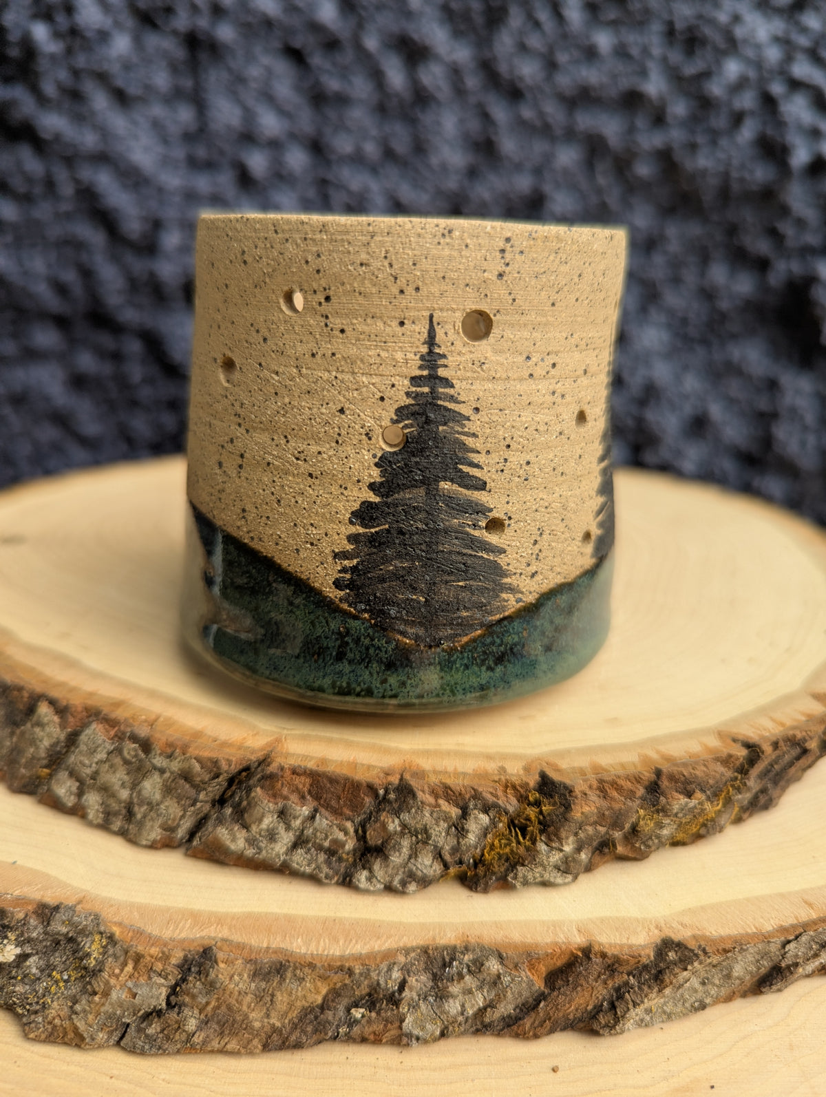 A handmade ceramic cup with a natural sandy beige body, featuring a hand-painted black pine tree design and a dark green glazed base, displayed on a rustic wood slice against a dark textured background.