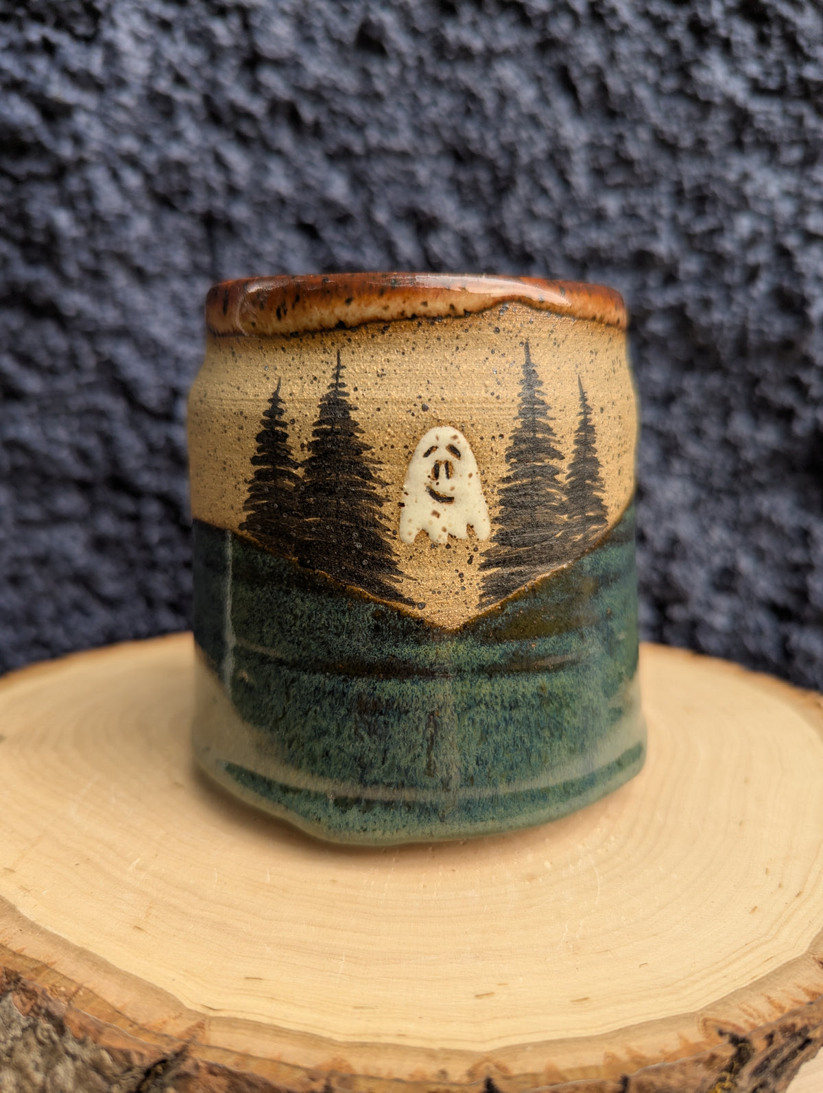 A handmade ceramic cup with a rustic landscape design, featuring a beige and green glaze with a carved or painted ghost and pine tree motif, displayed on a wood slice against a dark textured background.