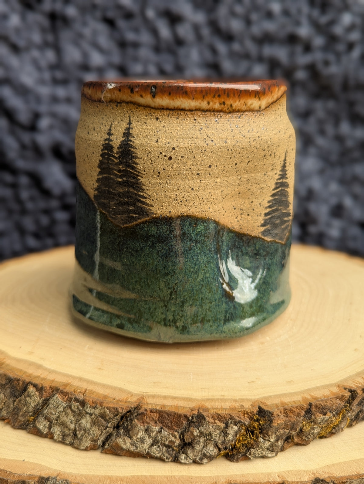 A handmade ceramic cup with a rustic landscape design, featuring pine trees and a white bird on a green and beige glazed background, displayed on a wood slice against a dark textured backdrop.