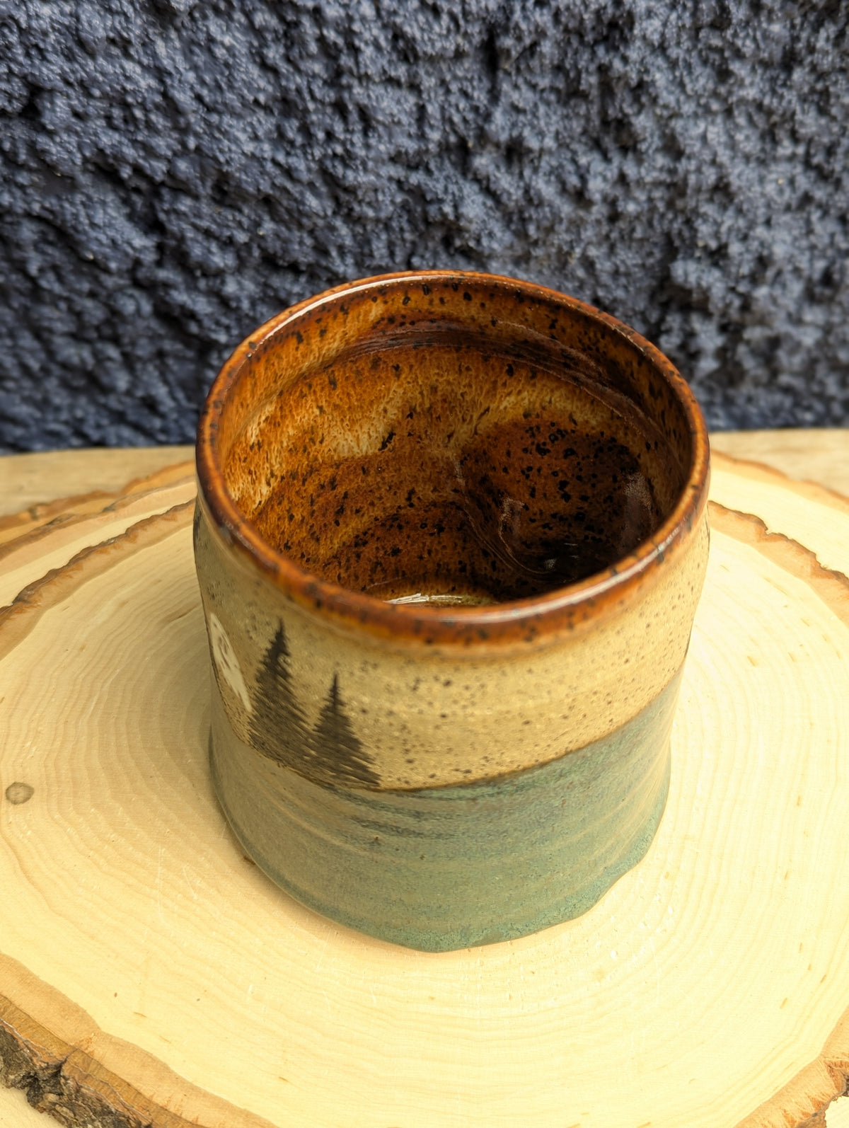 A handmade ceramic mug with a rustic brown and beige glaze and a painted pine tree design, sitting on a wooden slice coaster against a dark textured background.
