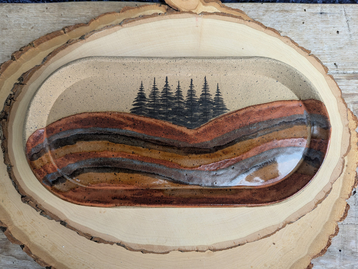 An oval ceramic tray with a glossy, wavy striped pattern in earthy tones of brown, terracotta, black, and beige, displayed on a rustic wood slice over a light wood background.