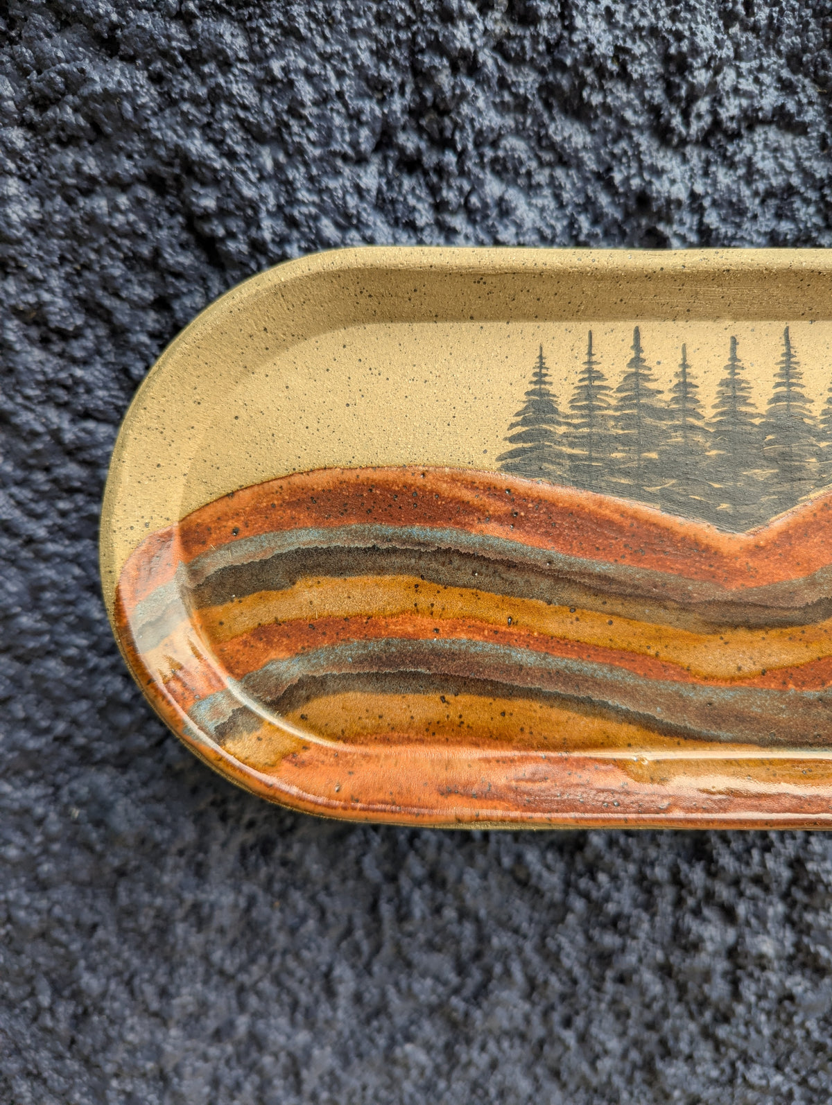 A close-up of a ceramic tray with rounded corners, featuring a landscape design of wavy orange, brown, and gray hills under a beige sky with black pine trees. The tray is placed on a dark gray textured fabric background.