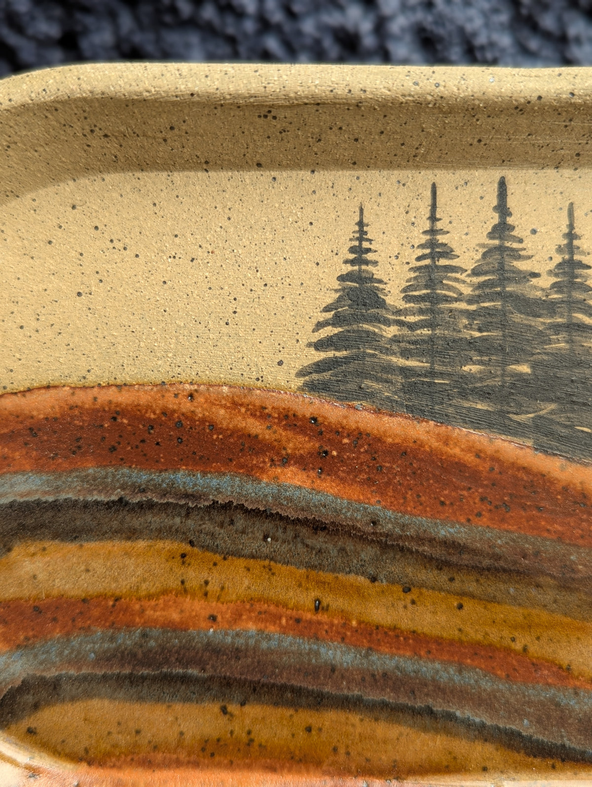 Close-up of a handmade ceramic tray featuring a hand-painted landscape design with pine trees under a speckled beige sky and layered earthy hills in orange, brown, and gray.
