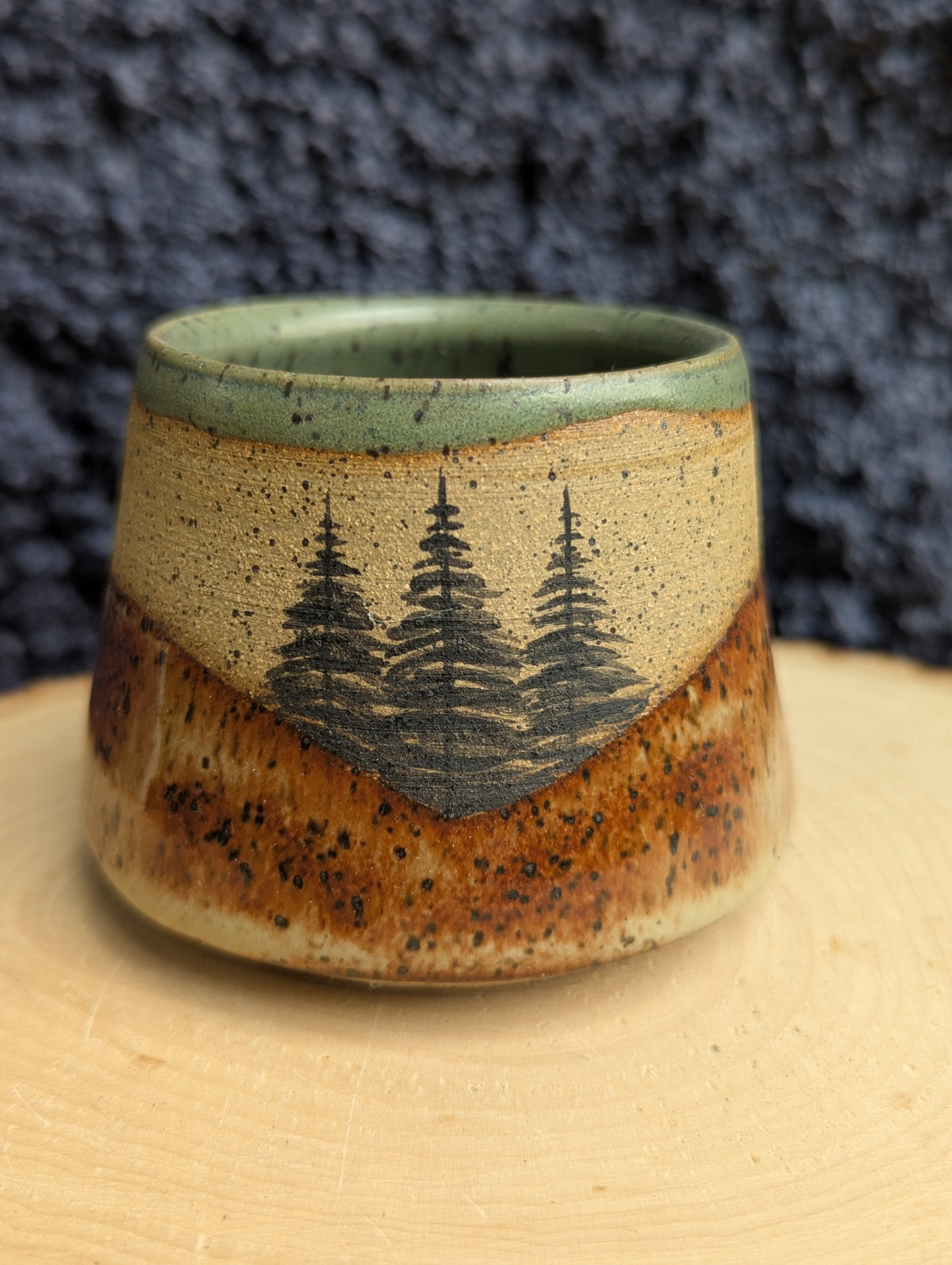 A handmade ceramic cup with a rustic, tapered shape, featuring a hand-painted design of three pine trees on a beige and brown speckled glaze, with a green rim, displayed on a wooden slice against a dark textured background.