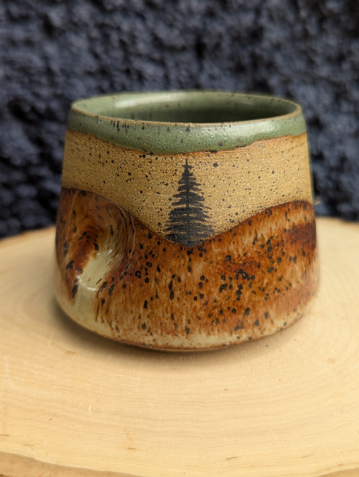 A handmade ceramic cup with a rustic design, featuring a speckled brown and beige body, a dark tree motif, and a green glazed rim, set on a wooden slice against a dark textured background.