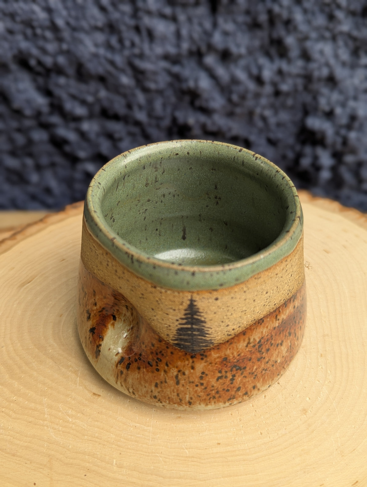 A handmade ceramic cup with a speckled green interior and a brown glazed exterior featuring a small pine tree design, displayed on a wooden slice with a dark textured background.