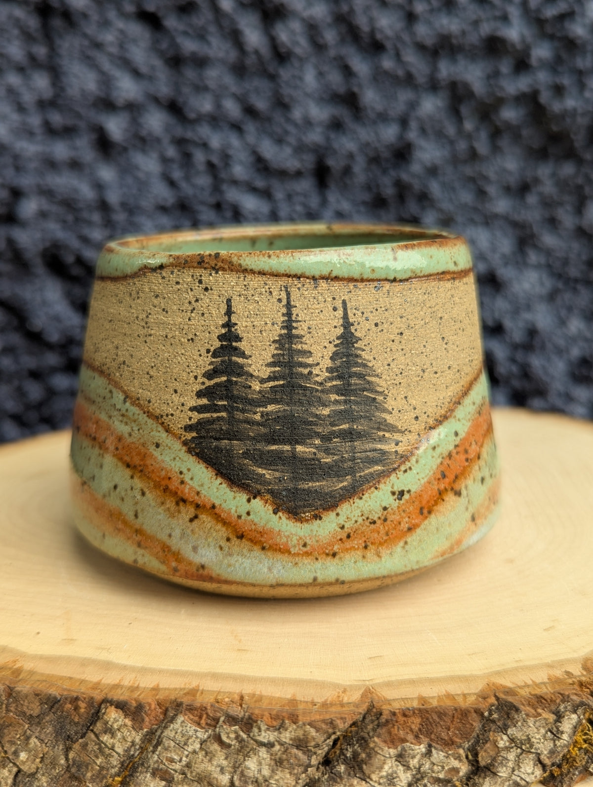 A handmade ceramic cup with a natural wood grain pattern and a black pine tree design, displayed on a rustic wood slice against a dark textured background.