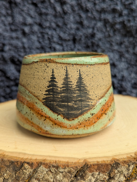 A handmade ceramic cup with a natural wood grain pattern and a black pine tree design, displayed on a rustic wood slice against a dark textured background.