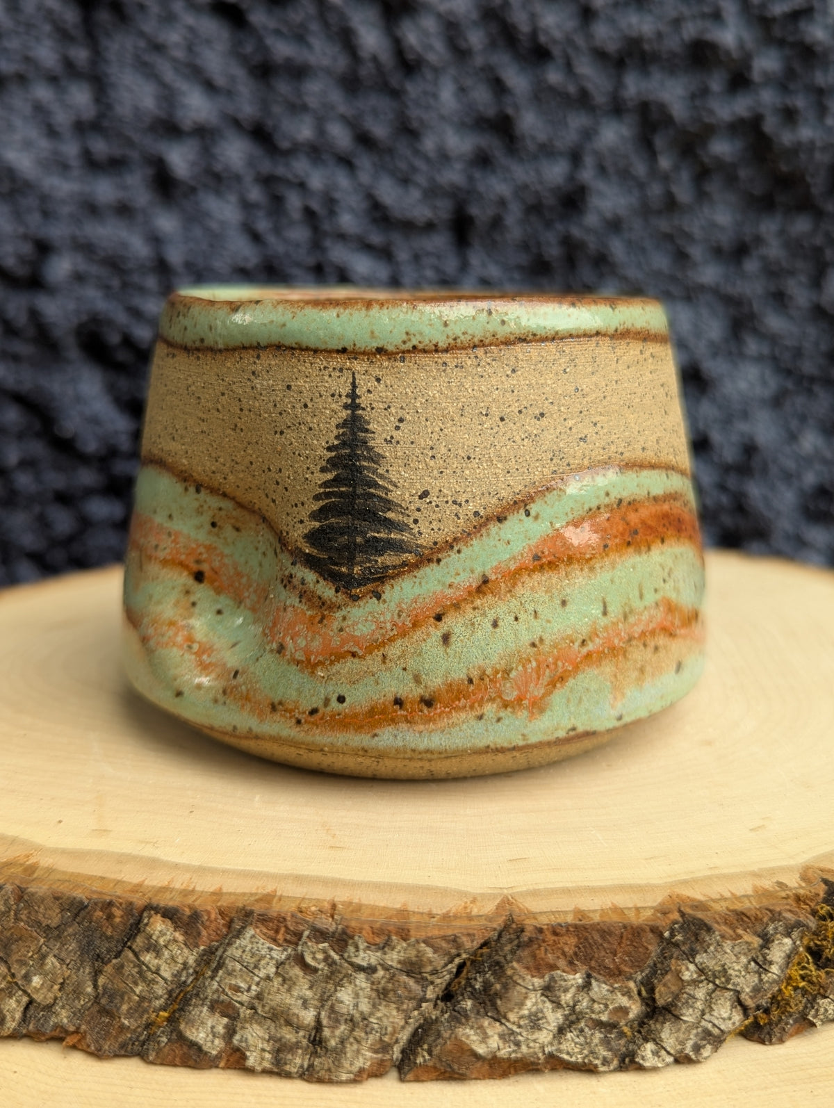 A handmade ceramic cup with a natural stoneware base, decorated with a light green glaze, orange-brown wavy stripes, and a black painted pine tree. The cup is displayed on a rustic wood slice against a dark textured background.
