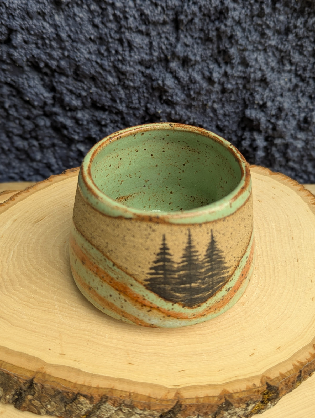 A handmade ceramic cup with a speckled beige and light green glaze, featuring a black printed pine tree design, displayed on a rustic wood slice against a dark textured background.