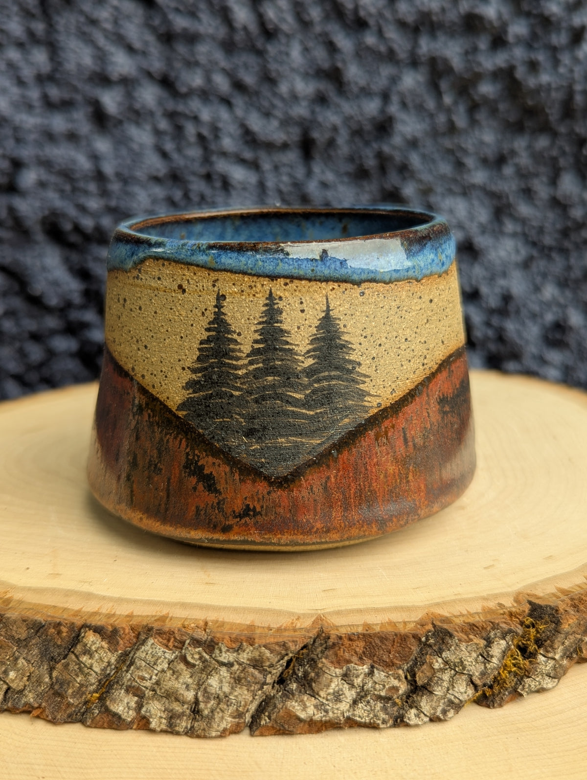 A handmade ceramic cup with a rustic mountain landscape design, featuring pine trees and a blue sky glaze, displayed on a wood slice against a dark textured background.