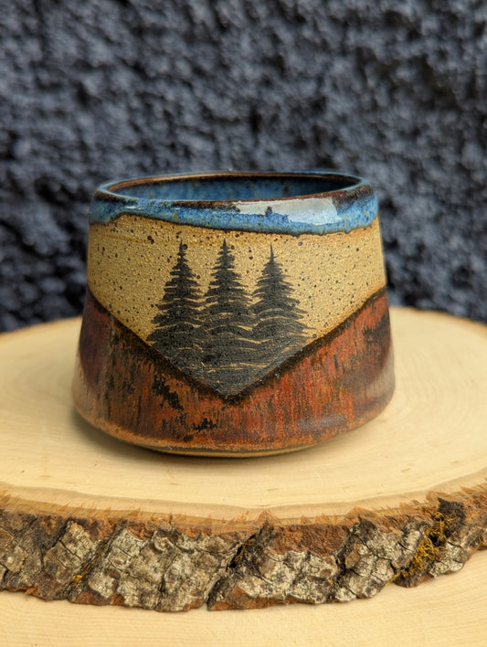 A handmade ceramic cup with a rustic mountain landscape design, featuring pine trees and a blue sky glaze, displayed on a wood slice against a dark textured background.