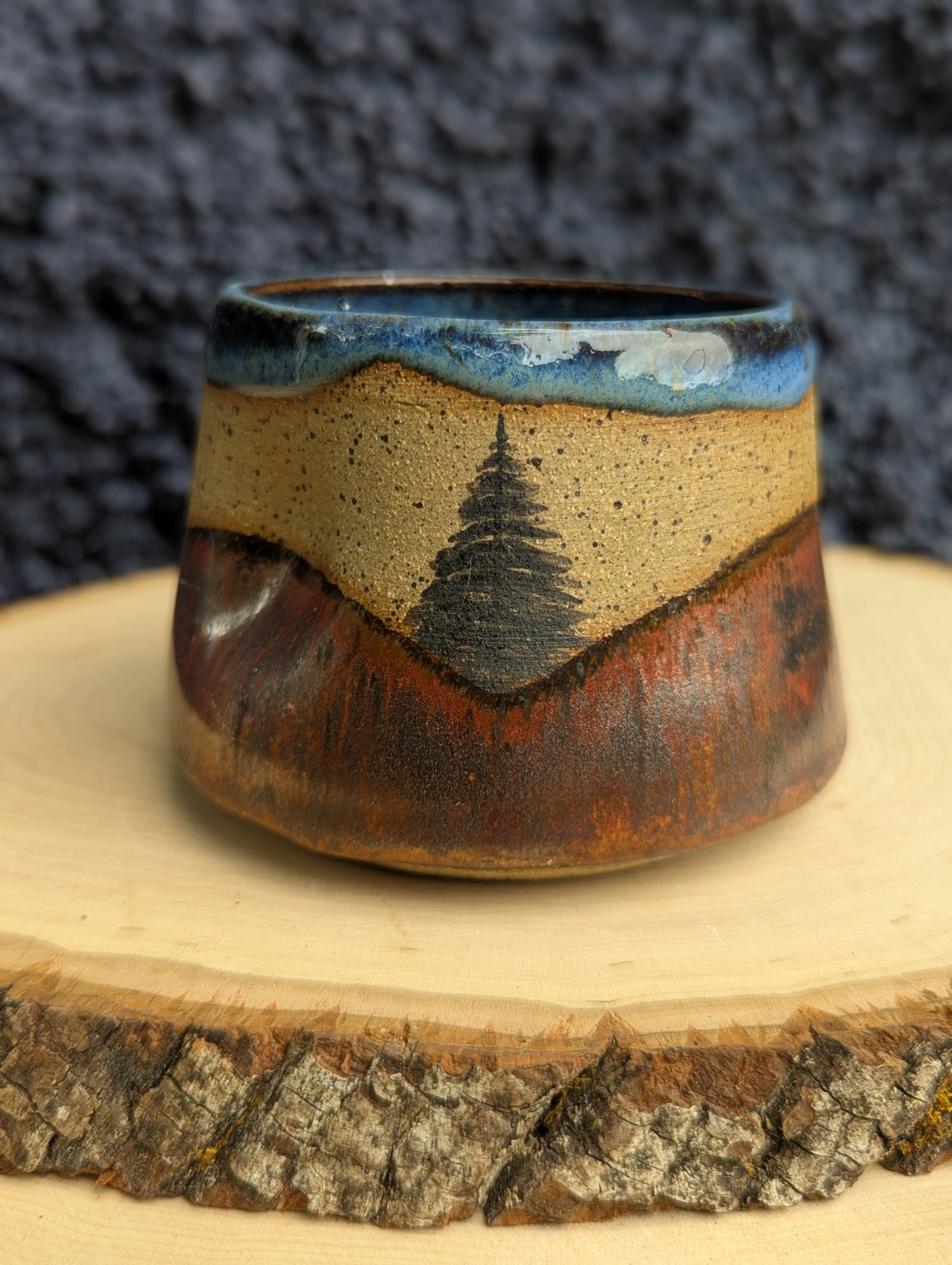 A handmade ceramic cup with a rustic mountain landscape design, featuring a blue sky, beige mountains, and a pine tree, displayed on a wood slice against a dark textured background.