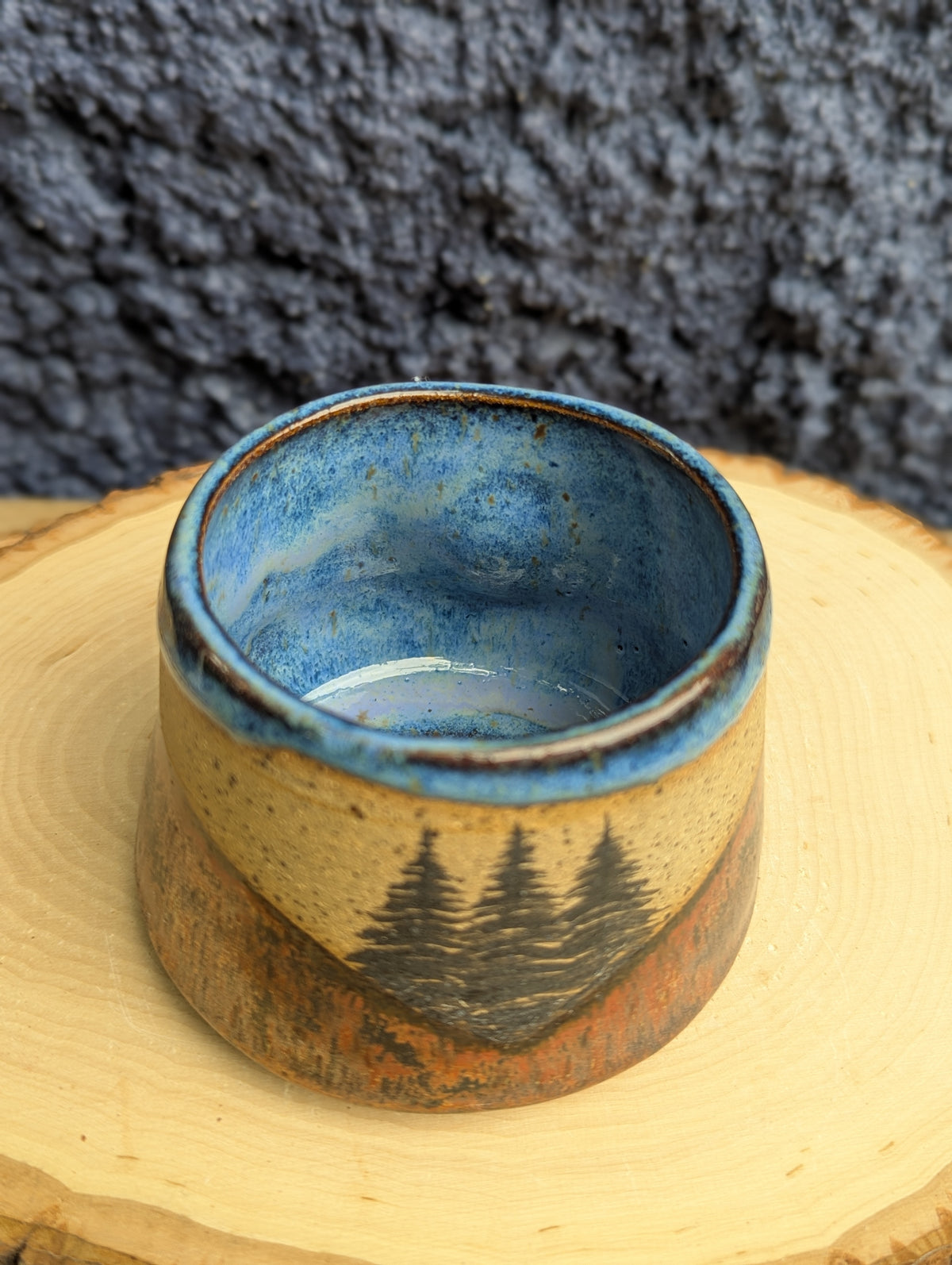 A handmade ceramic cup with a blue glazed interior and a rustic tan exterior featuring a painted pine tree design, displayed on a wood slice with a dark textured background.